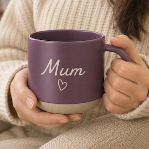 Op de afbeelding: Een paarse keramische mok met een matte afwerking, voorzien van het woord Mum in witte schrijfletters boven een kleine witte hartvorm. De mok heeft een glad bovenste gedeelte en een textuurrijke, lichtbeige onderkant met een gebogen oor.