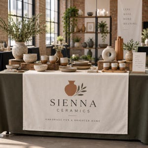 May include: A display table covered with a cream-coloured cloth featuring the Sienna Ceramics logo and the text 'Handmade for a brighter home'. The table is arranged with various ceramic bowls, mugs, vases, and plates in neutral, earthy tones.