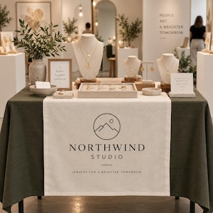 May include: A display table covered with a dark green cloth and a central beige runner featuring the Northwind Studio logo and text. The table holds jewellery on beige busts, a ring tray, small bowls, and framed signs with text about a brighter tomorrow.