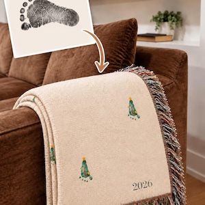 May include: A cream-colored woven throw blanket featuring a repeating pattern of green Christmas tree graphics that resemble baby footprints, topped with yellow stars. The blanket includes the year 2026 printed in black and has multi-colored fringe.