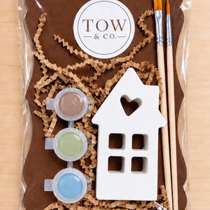 May include: A craft kit in a clear plastic bag containing a white wooden house cutout with a heart-shaped window, two paintbrushes with light wood handles, and three small circular paint pots in brown, green, and blue, with a TOW & CO. label.