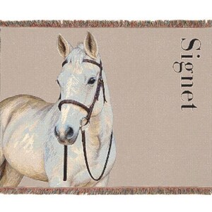 May include: A woven throw blanket featuring a light-colored horse wearing a dark brown bridle. The background is a solid tan color with the word Signet printed vertically in black on the right side. The blanket has a fringed edge.