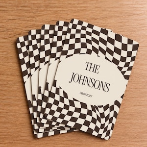 May include: A stack of rectangular cards featuring a black and white wavy checkerboard pattern. A central cream-colored oval contains the black text THE JOHNSONS and the date 08.07.2027 printed in a serif font. The cards have rounded corners.