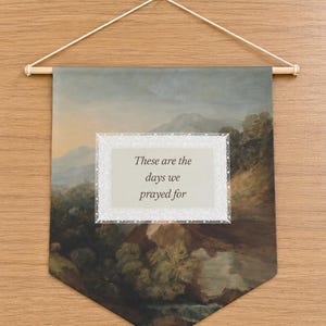 May include: A rectangular fabric wall hanging featuring a landscape painting of rolling hills and trees. A white rectangular inset with a lace-like border contains the black text: These are the days we prayed for. The banner hangs from a wooden rod.