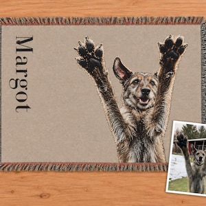 A rectangular woven throw blanket featuring a tan background with a fringed edge. The blanket displays a large, detailed image of a dog with its front paws raised in the air and the name Margot printed in black vertical text. Dog memorial blanket