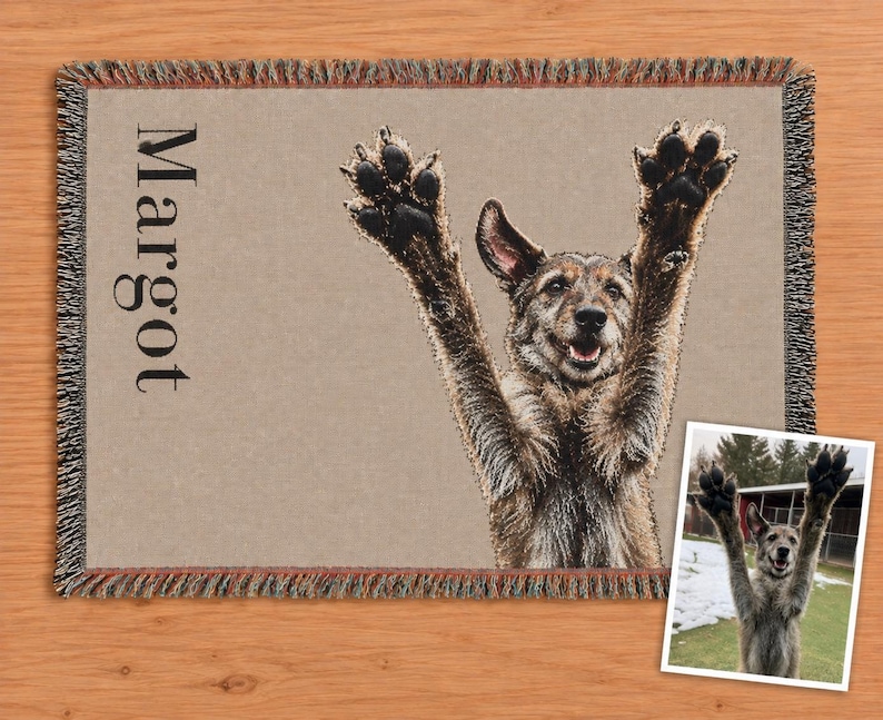 A rectangular woven throw blanket featuring a tan background with a fringed edge. The blanket displays a large, detailed image of a dog with its front paws raised in the air and the name Margot printed in black vertical text. Dog memorial blanket
