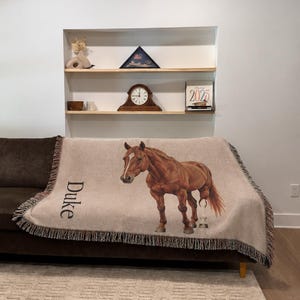A woven throw blanket in a light beige color featuring a realistic illustration of a brown horse and the name Duke printed in black text. The blanket has a fringed edge and is draped over a dark brown sofa in a living room setting. Custom horse blanket