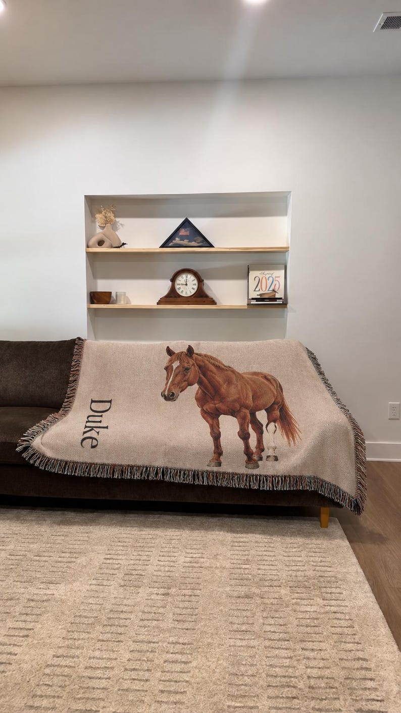 A woven throw blanket in a light beige color featuring a realistic illustration of a brown horse and the name Duke printed in black text. The blanket has a fringed edge and is draped over a dark brown sofa in a living room setting. Custom horse blanket