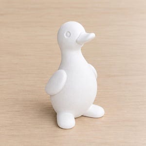 May include: A small, matte white duck figurine with a smooth, rounded body, a simple beak, and defined circular eyes. The duck stands in an upright position with its wings tucked against its sides and feet pointing forward, featuring a clean finish.