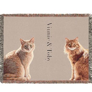 May include: A rectangular woven throw blanket with a light beige background featuring two orange cats sitting on either side of the text Vinnie & Toby. The blanket has a fringed edge with multi-colored threads along all four sides.