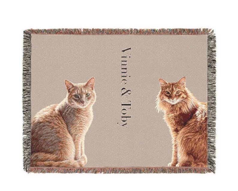 May include: A rectangular woven throw blanket with a light beige background featuring two orange cats sitting on either side of the text Vinnie & Toby. The blanket has a fringed edge with multi-colored threads along all four sides.