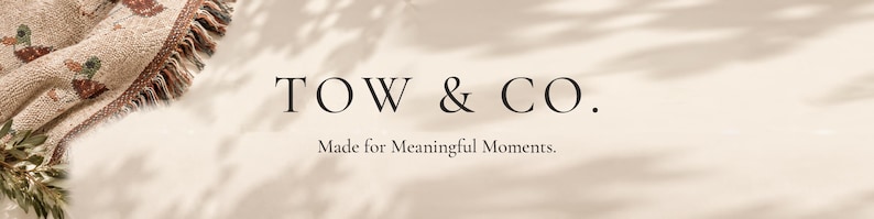 May include: A woven blanket with a duck pattern and fringed edges, partially visible in the corner. The text TOW & CO. and Made for Meaningful Moments. is centered on a light beige background with soft, dappled leaf shadows.