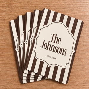 May include: A stack of rectangular cards featuring a vertical brown and cream striped pattern. Each card displays a central cream-colored decorative frame containing the text The Johnsons and the date 26.06.2026 in a dark brown serif font.