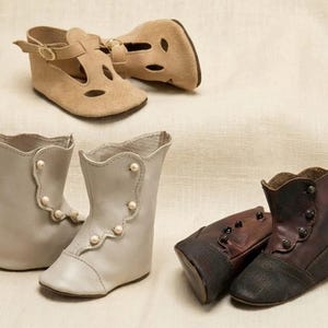 May include: A collection of footwear including a pair of tan suede T-strap shoes with gold buckles, a pair of light grey leather boots with pearl-like buttons along the side, and a pair of dark brown leather boots with dark buttons.