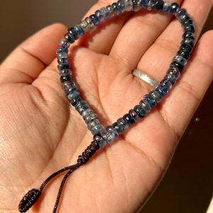 May include: A bracelet composed of small, translucent, round blue beads with varying shades of deep navy and light sky blue. The bracelet features a dark brown adjustable cord closure with two small knotted ends hanging from the main circular band.
