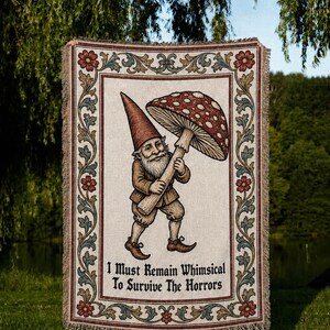 Puede incluir: Una manta tejida con una ilustración central de un gnomo sosteniendo una seta grande roja con puntos blancos. El texto "I Must Remain Whimsical To Survive The Horrors" está impreso debajo del gnomo, enmarcado por un borde floral.
