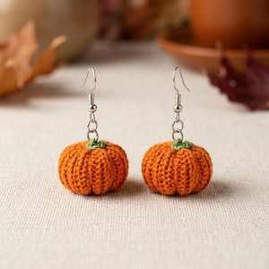 May include: A pair of dangle earrings featuring small, crocheted orange pumpkins with tiny green stems. Each pumpkin is attached to a silver-toned metal fishhook ear wire by a short silver chain link, creating a festive autumn-themed accessory.