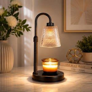 May include: A black metal candle warmer lamp with a curved arm and a clear, textured glass shade. The lamp features a round base holding a small glass jar candle with a glowing, lit wick, providing warm illumination to the surrounding area.