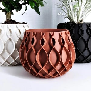 Op de afbeelding: Een terracotta-kleurige plantenpot met een herhalend geometrisch rasterpatroon aan de buitenkant. De pot is rond met een brede opening en staat voor twee andere soortgelijke potten, één in het wit en één in het zwart, die elk een plant bevatten.