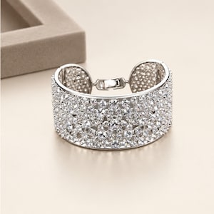 Può includere: Un ampio bracciale a fascia color argento caratterizzato da una disposizione densa e complessa di pietre trasparenti sfaccettate che coprono l'intera superficie esterna. Il bracciale include un meccanismo di chiusura in metallo.