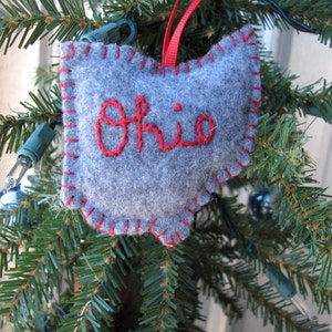 May include: A gray felt Ohio state ornament with red stitching and the word "Ohio" embroidered in red. The ornament hangs from a red ribbon on a Christmas tree with green needles and small lights.