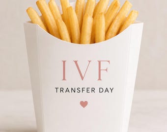 Puede incluir: Un recipiente de papel blanco lleno de patatas fritas doradas. La parte frontal del recipiente presenta el texto "IVF" en letras rosas grandes, "TRANSFER DAY" en letras negras más pequeñas y un pequeño icono de corazón rosa bajo el texto.