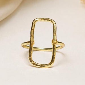 May include: A gold ring featuring a vertical, rectangular-shaped open frame. The metal surface of the rectangular element has a textured, hammered finish, while the circular band remains smooth and polished in appearance.