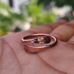 May include: A copper-colored metal ring with an open, bypass-style design. The band features a hammered, textured finish with flattened, rounded ends that overlap slightly. The surface of the metal displays a warm, metallic reddish-brown tone.