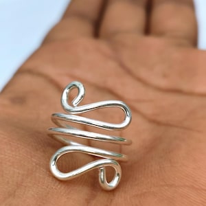 May include: A silver-coloured ring crafted from a single continuous wire that forms a series of stacked, open loops. The design features a spiral-like structure with rounded ends, creating a fluid, interconnected pattern that wraps around the finger.
