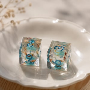 Könnte beinhalten: Zwei würfelförmige Spielwürfel aus klarem Kunstharz mit eingebetteten blauen und orangefarbenen floralen Elementen. Jeder Würfel hat abgerundete Kanten und vertiefte, metallisch blau bemalte Zahlen, wobei die Ziffern zwei, neun, eins und drei sichtbar sind.
