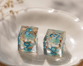 May include: Two clear resin cube-shaped dice featuring embedded blue and orange floral elements. Each die has rounded edges and recessed numbers painted in a metallic blue finish, with the numbers two, nine, one, and three clearly visible.