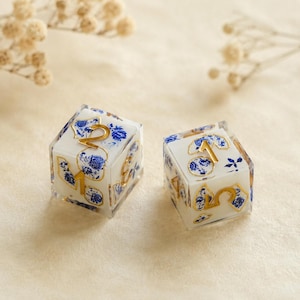May include: Two six-sided dice featuring a white base with a blue floral pattern and gold-colored numbers. The dice are crafted from a clear material, showcasing the intricate blue and white botanical design on each face with metallic gold numerals.