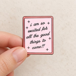 May include: A rectangular enamel pin with a light pink background and a dark pink border. The pin features black cursive text that reads, "i am so excited for all the good things to come!!!" accented by small black plus signs.