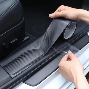 Könnte beinhalten: Eine Rolle schwarze Klebefolie mit Carbonfaser-Struktur wird auf die Türschwelle eines Fahrzeugs aufgebracht. Die Folie weist ein ausgeprägtes diagonales Webmuster und eine glänzende Oberfläche auf, wobei eine transparente Schutzfolie abgezogen wird.