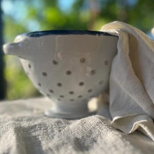 Könnte beinhalten: Ein weißes Keramik-Sieb mit blauem Rand und kleinen kreisförmigen Abflusslöchern in der Schale. Das Sieb verfügt über zwei kleine seitliche Griffe und einen Standfuß, begleitet von einem gefalteten, cremeweißen Leinentuch an der Seite.