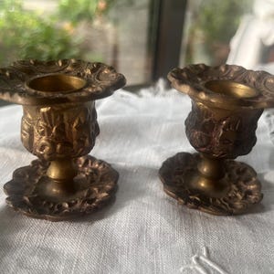 Puede incluir: Dos candelabros de latón a juego con un borde ancho y festoneado y una base circular. La columna central y la base están decoradas con intrincados patrones florales y de volutas en relieve, con un acabado en tono dorado metálico envejecido.