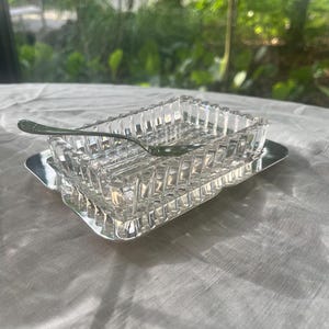 May include: A rectangular clear glass butter dish with a textured, faceted geometric pattern on the sides. The dish rests on a polished silver-toned metal tray and includes a small, matching silver-toned metal butter knife resting across the top.