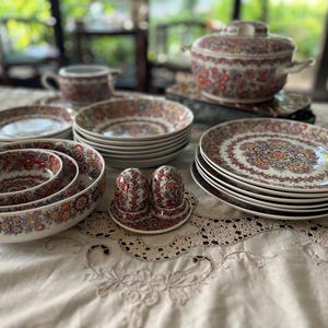 Może przedstawiać: Zestaw ceramicznej zastawy stołowej na białym tle z gęstymi, kwiatowymi i geometrycznymi wzorami w odcieniach czerwieni, pomarańczu, błękitu i złota. Komplet zawiera stosy talerzy obiadowych, miski, wazę z pokrywką, dzbanek na śmietankę oraz solniczkę i pieprzniczkę.