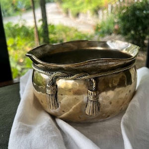 Pode incluir: Uma taça redonda de latão martelado com um acabamento decorativo de corda em torno da parte superior, acentuada com duas borlas pendentes. A taça tem uma borda larga, ondulada e irregular, com um acabamento metálico polido que reflete a luz.