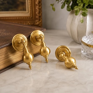 May include: Three brass drawer pulls with a brushed gold finish. Each pull features a circular mounting plate and a teardrop-shaped pendant handle that hangs from a central hinge. The hardware has a smooth, metallic surface and a pointed tip.
