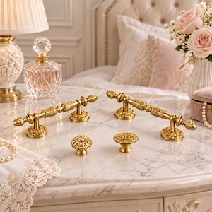 May include: Two gold-toned metal drawer pulls with ornate, textured detailing and two matching round cabinet knobs featuring a floral-inspired pattern. The hardware pieces have a polished finish and are arranged on a white marble surface.