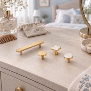 May include: A collection of cabinet hardware including one long rectangular gold handle, one flat circular gold knob, one clover-shaped gold knob with a white inlay, and one circular gold knob with a white inlay, all arranged on a light wood surface.
