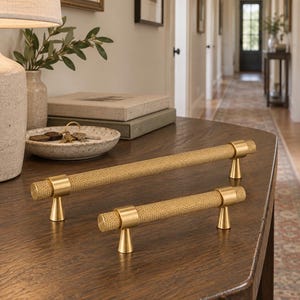 May include: Two gold-toned metal cabinet pulls with a textured, knurled cylindrical bar and smooth, conical mounting posts. The hardware features a metallic finish and a repeating cross-hatch pattern along the length of each handle.