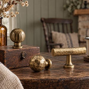 May include: Three brass cabinet knobs with a hammered texture. Two knobs are spherical in shape, while one knob is a horizontal T-bar design. Each piece features a brushed metallic finish with visible indentations across the surface.