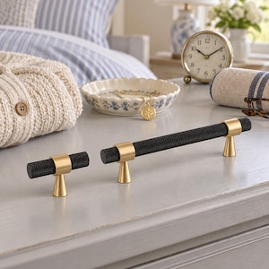 May include: Two cabinet pulls featuring a textured black cylindrical bar with a knurled finish, supported by two smooth, conical brass-toned metal mounts. The hardware displays a contrasting combination of matte black and polished gold-toned metal.