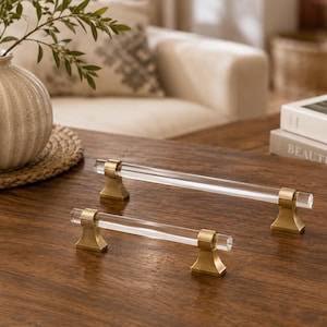 May include: Two cabinet pulls featuring clear cylindrical acrylic bars held by brushed brass metal mounts. The hardware consists of one longer pull and one shorter pull, each designed with two sturdy, flared pedestal bases for attachment.