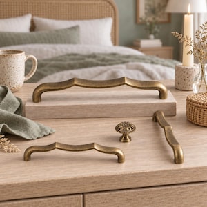 May include: A collection of antique brass hardware including two long arched cabinet pulls, one smaller arched cabinet pull, and one round floral-patterned cabinet knob. The hardware features a weathered metallic finish with curved silhouettes.