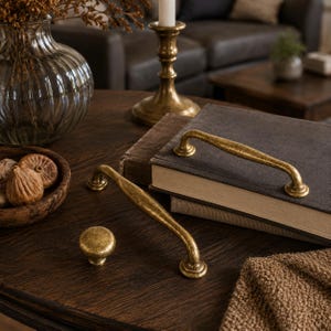 May include: Two curved metal drawer pulls and one round cabinet knob, all featuring a distressed, antique gold finish with a textured surface. The hardware pieces are arranged on a dark wooden surface alongside a stack of books and a glass vase.