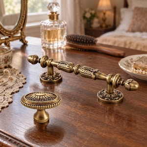 Puede incluir: Un tirador de mueble de metal en tono latón con una barra cilíndrica alargada y ornamentada y dos bases de montaje circulares, acompañado de un pomo de mueble redondo a juego con un patrón floral texturizado y detallado.