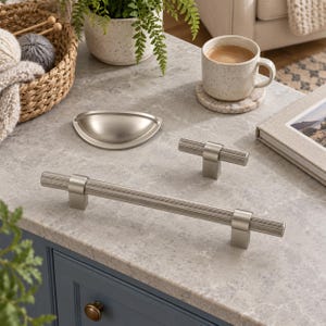 May include: A collection of brushed nickel cabinet hardware including one long cylindrical bar pull with a ribbed texture, one smaller matching T-shaped knob, and one semi-circular cup pull, all featuring a smooth metallic finish.